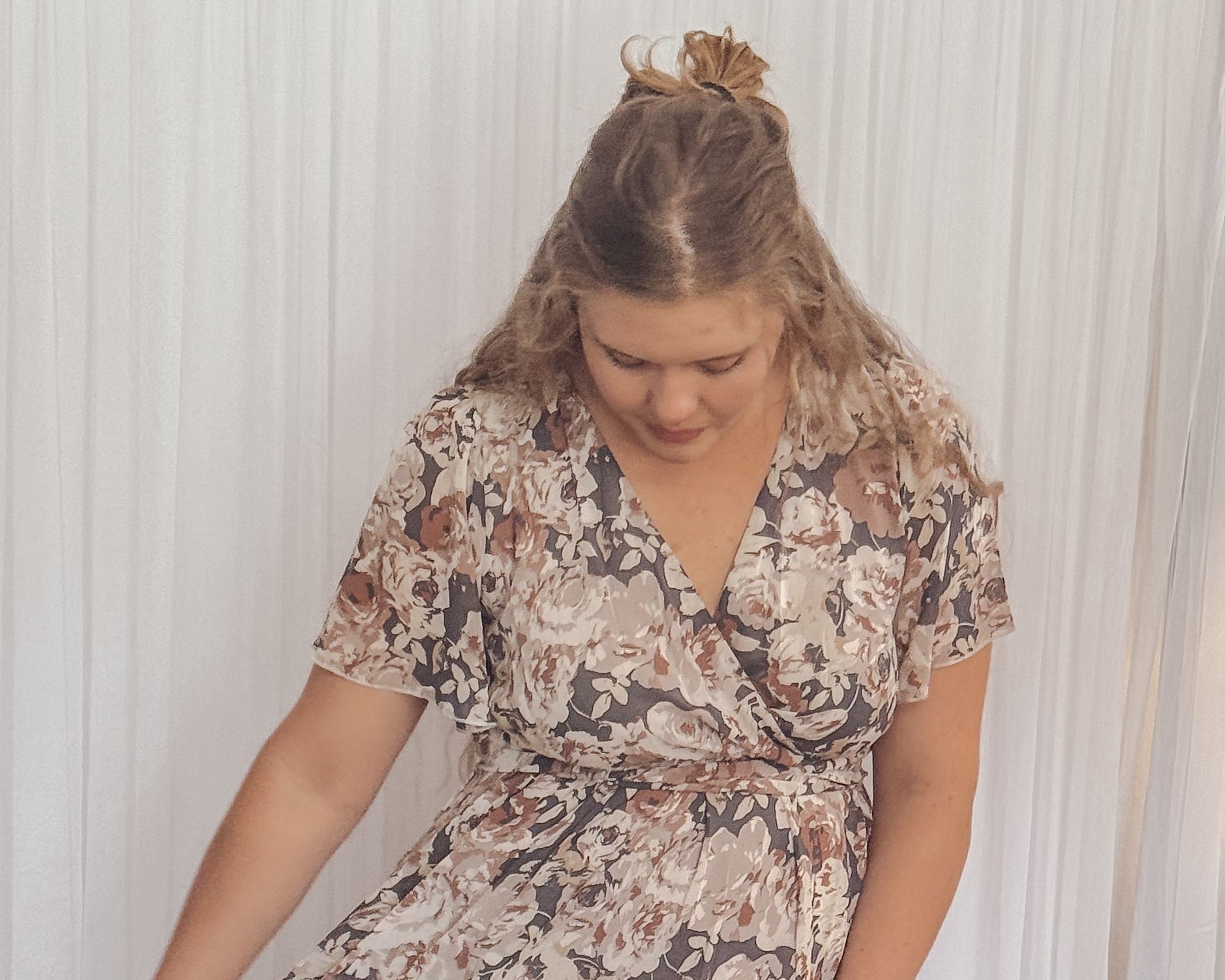 Woman wearing a floral dress standing in a room with white curtains and a nightstand.